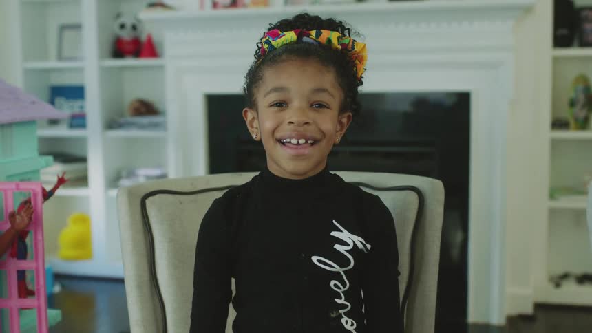 A pretty little girl smiles while in a chair near toys and the fireplace
