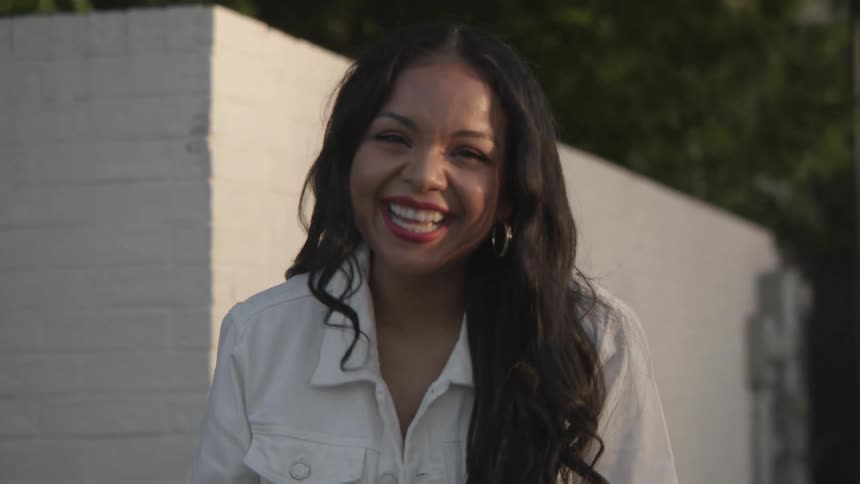 A woman laughs and smiles as she poses outside a white brick building