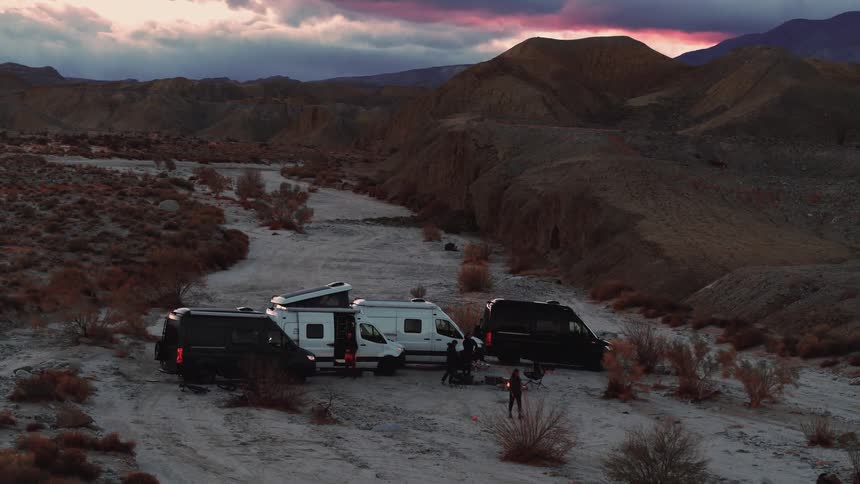 A group of people stand around a fire by their vehicles at the base of a canyon as one woman walks after a dog