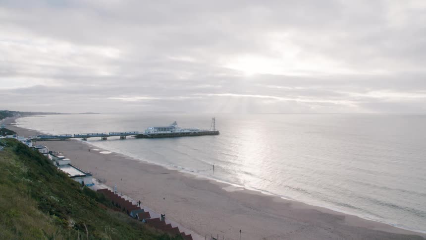 Person walks across beach near shed as bird flies over head and wave roll in past dock