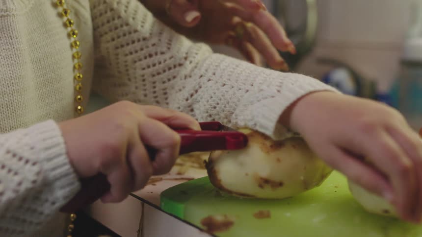 Middle aged woman with rings helps girl with white sweater peeling potatoes on cutting board in the kitchen