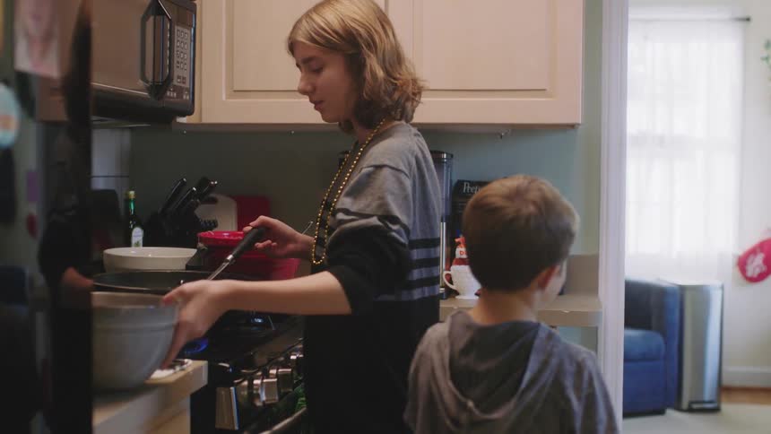 A boy hands a girl a bowl as she cooks at the stove