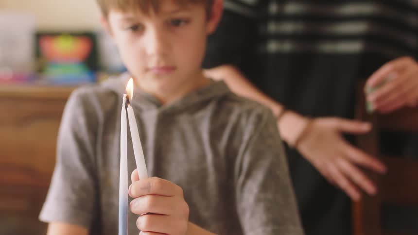 A young boy at home uses a candle to light another candle as his brother stands behind him and watches