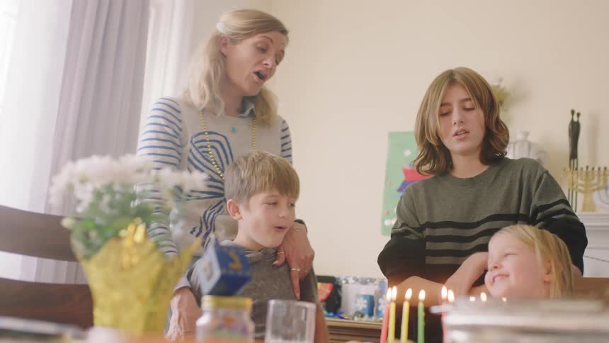 A little girl sits at a table in front of a cake with candles and laughs as her two brothers and mother sing to her