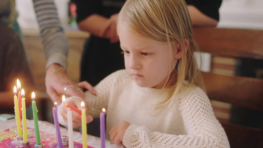 A young girl sits at home and lights a candle with another candle as her mother and brothers watch