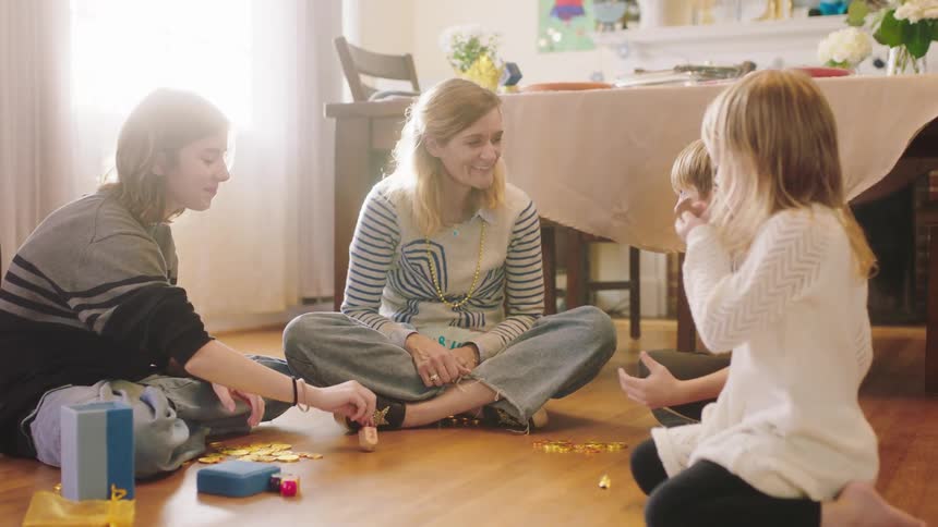 Mother with her son and daughters sitting on the floor at home playing with dreidels on hanukkah