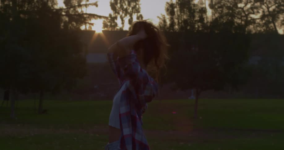 A teenager pulls her long hair up into a ponytail while standing in a park