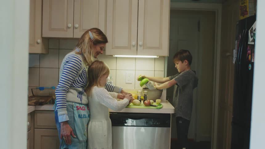 Mother stands in kitchen guiding daughter and son as they help her prepare a meal