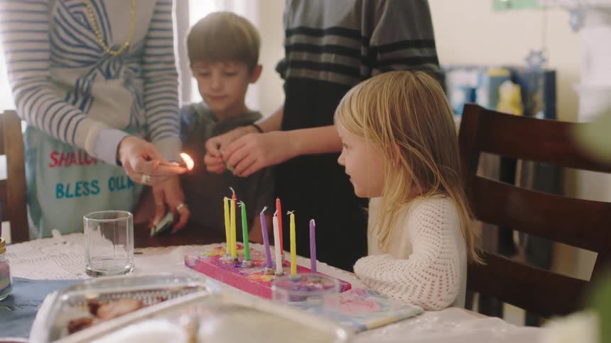 Mother lights the candles on a birthday cake in front of a small happy blonde girl at home