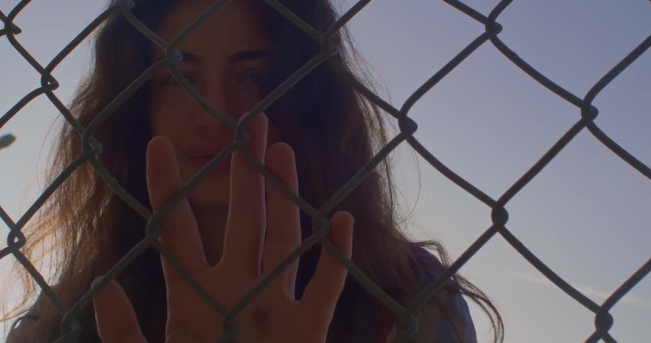 A teenage girl is holding her hand against a chain link fence and looking sad