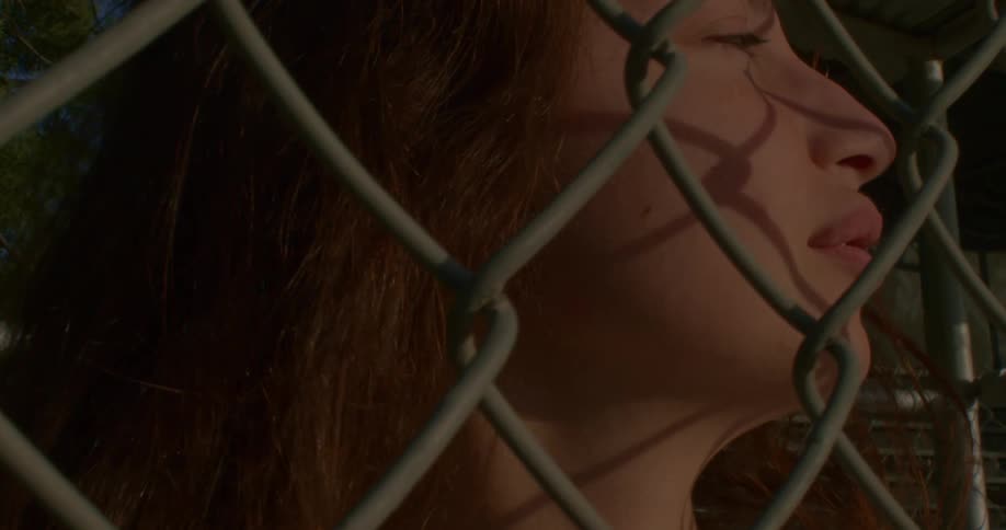 A girl leans against a chain link fence and looks off in the distance