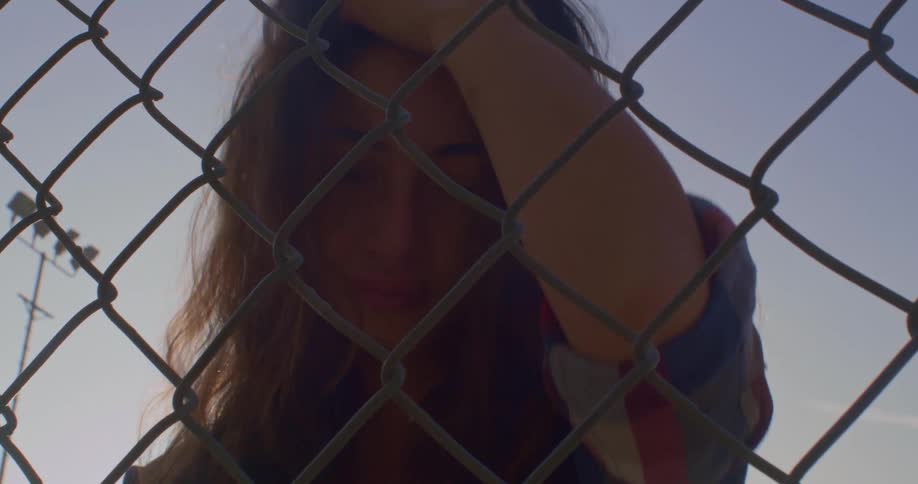 Young woman pulls her hair up with her hand standing in front of a wired fence at sunset