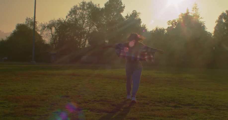 Woman with brown wavy hair flannel shirt and jeans walks with open arms as the sun shines brightly in the public park