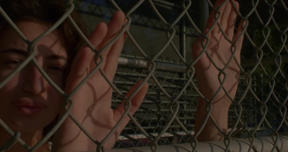 A woman leans up against a chain link fence and looks ahead