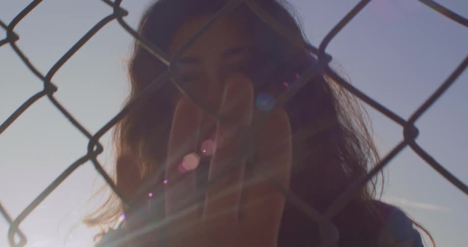 Young woman looking at the camera brushes her hand over a wired fence at sunset