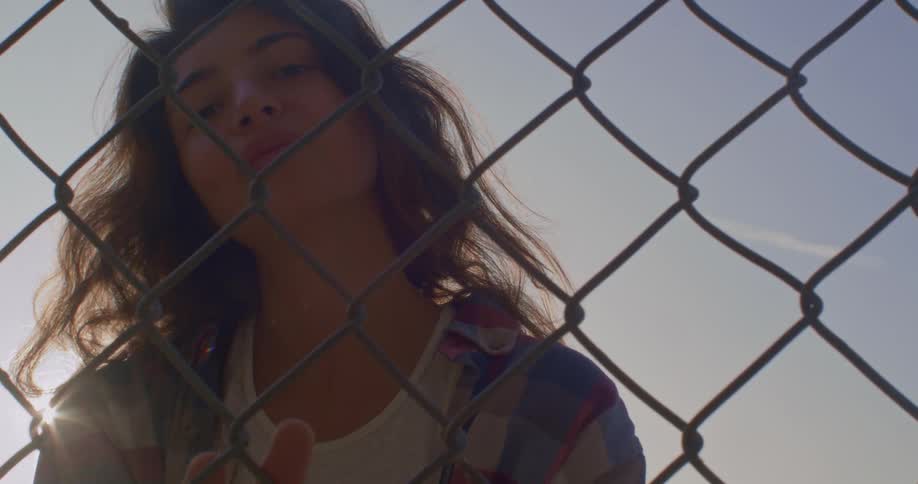 Young woman looking at the camera slides her hands over a wired fence at sunset