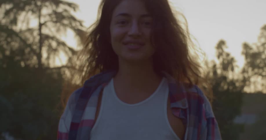 Woman with brown wavy hair and flannel shirt walks towards camera smiling in public park at dusk