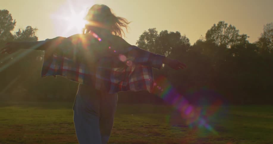 Young woman with arms wide open walking towards the sunset in a park