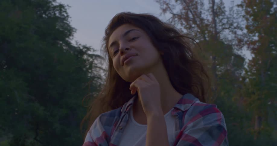Woman with brown curly hair flannel shirt and hand on chin faces the camera with eye contact smiling in public park at dusk