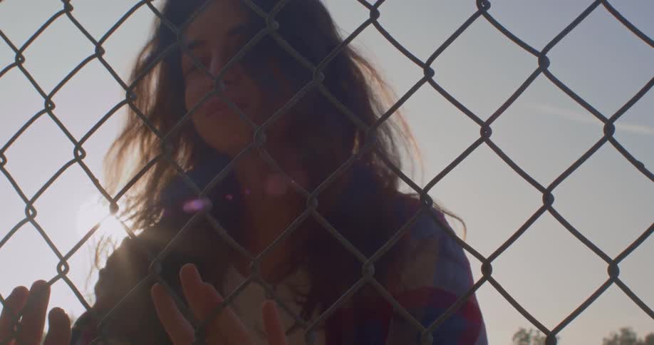 A young woman standing outdoors turns her head to look into the camera through a chain link fence