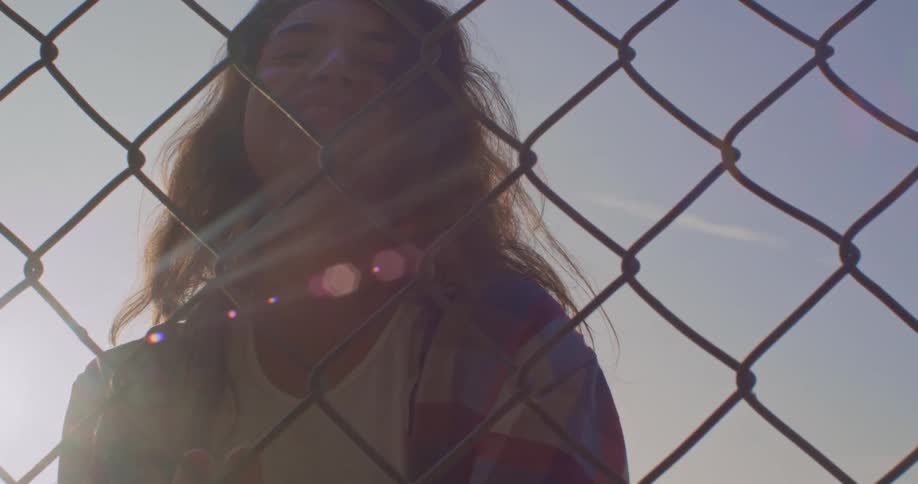 Young woman looking at the camera slides her hands over a wired fence at sunset