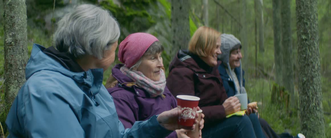 Older friends sit on forest slope with coffee cups talking and smiling