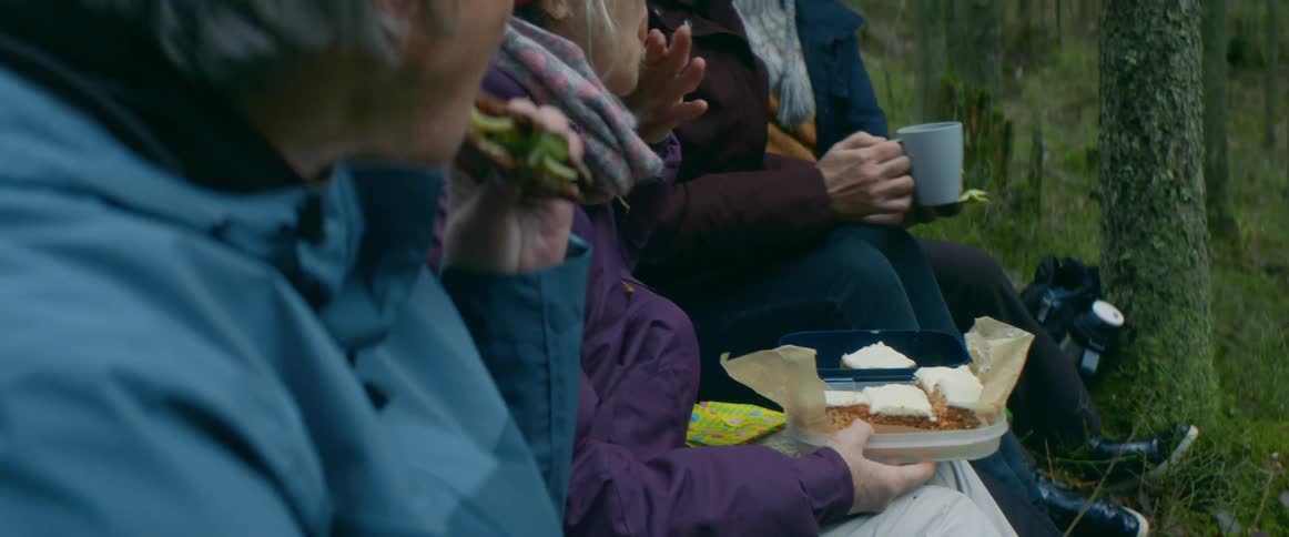 A group of elderly women wearing purple and blue jackets eats sandwiches and drinks from cups in the forest