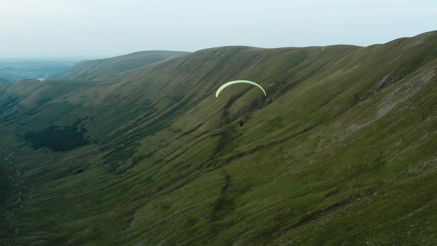 Person paraglides through green valley with mist covered horizon in background