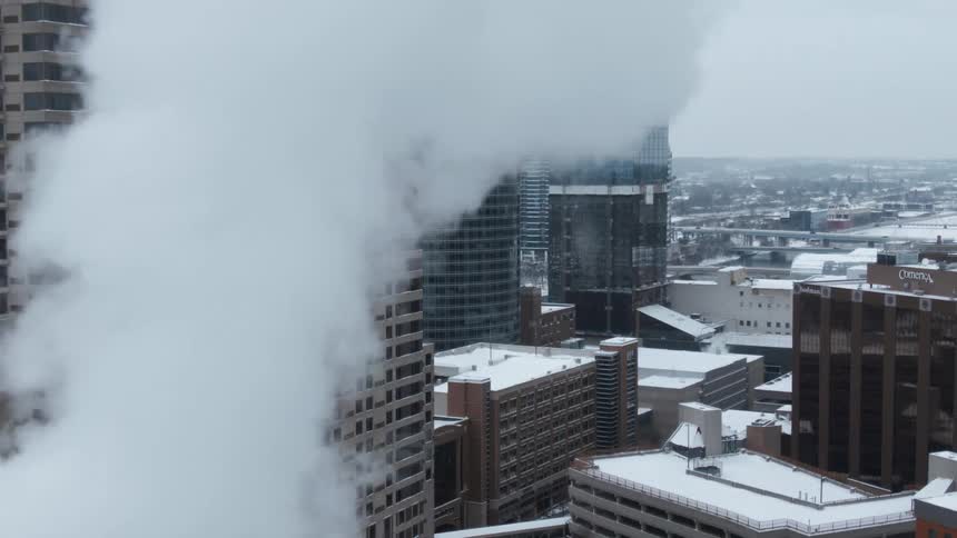 Vapor flows past snow topped skyscrapers as traffic flows across bridges in the misty background