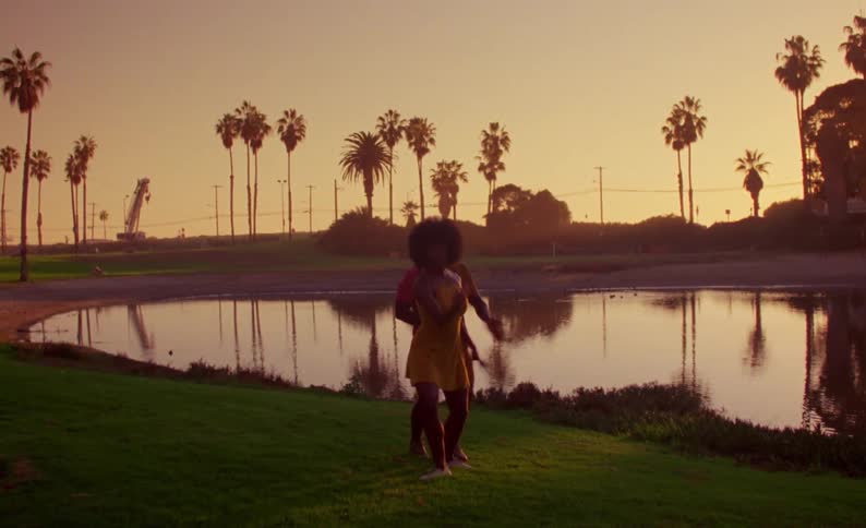 Female dancer with yellow dress and curly hair and male dancer with braids orange shirt and shorts dance on lake bank at sunset