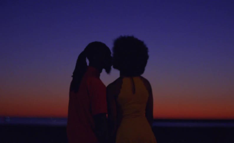 A man and woman turn to each other and kiss as they stand by the water at sunset
