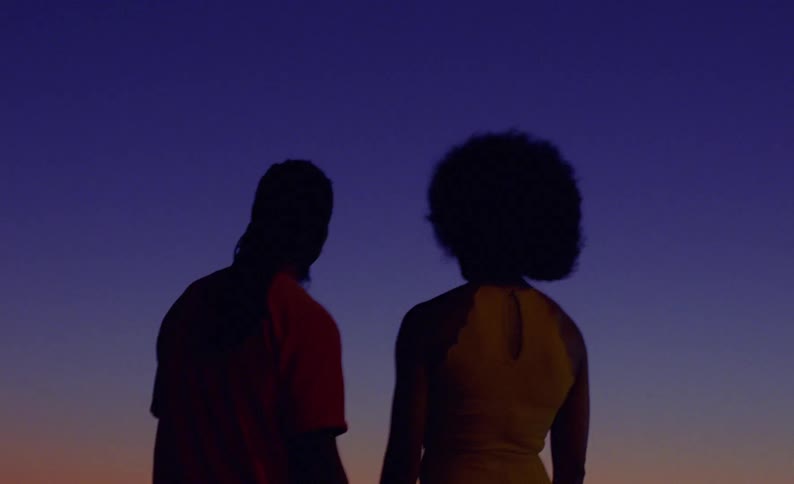 Couple stand on beach looking toward red tinted horizon and silhouetted against blue of the sky
