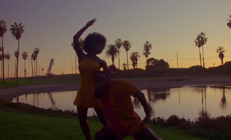 Male and female dancers dance on shore at park near pond under a red sky