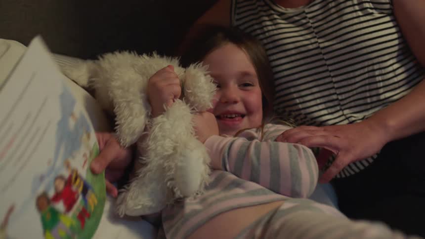 Mother with sleeveless shirt holds childrens book playing with her daughter on bed in bedroom