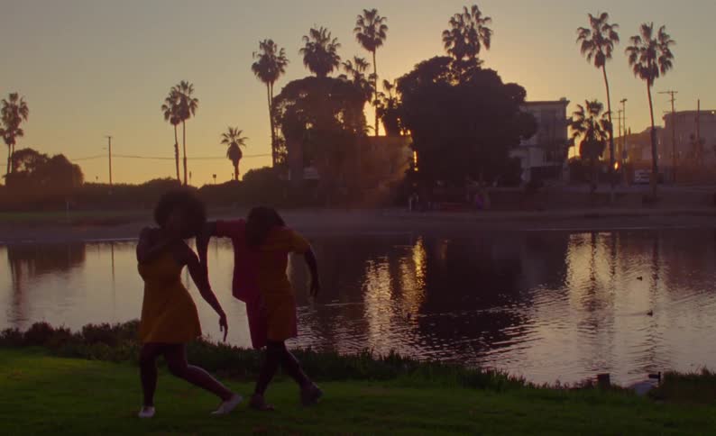 Male and female dancers dance on grassy shore near duck pond backed by red sky