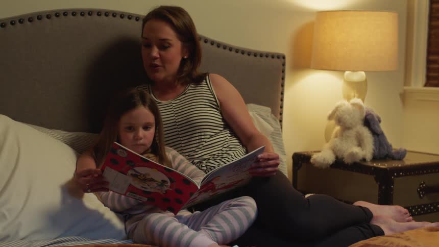 Mother with sleeveless shirt reads daughter book sitting on bed in bedroom