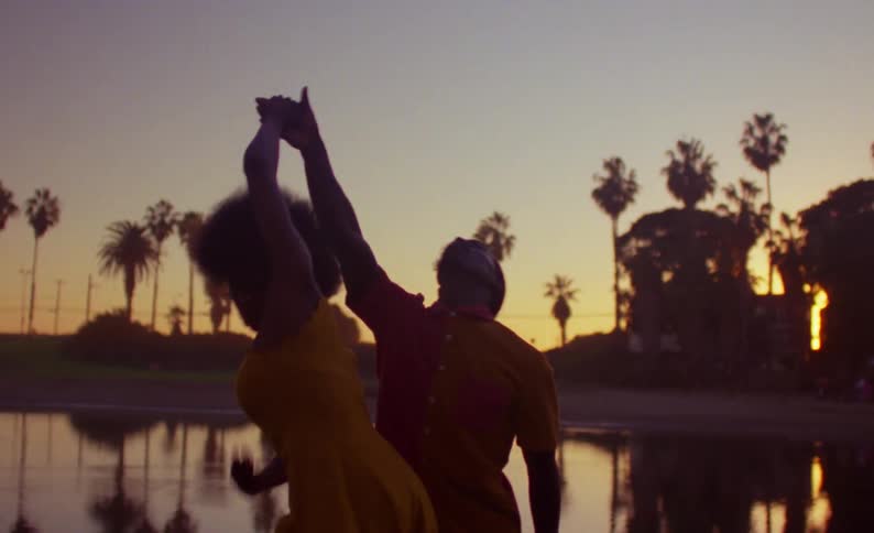 Female dancer with yellow dress and curly hair and male dancer with braids yellow shirt and shorts dance on lake bank at dusk