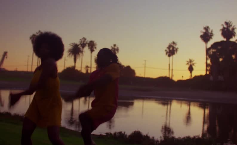 Female dancer with yellow dress and curly hair and male dancer with braids yellow shirt and shorts dance on lake bank at sunset