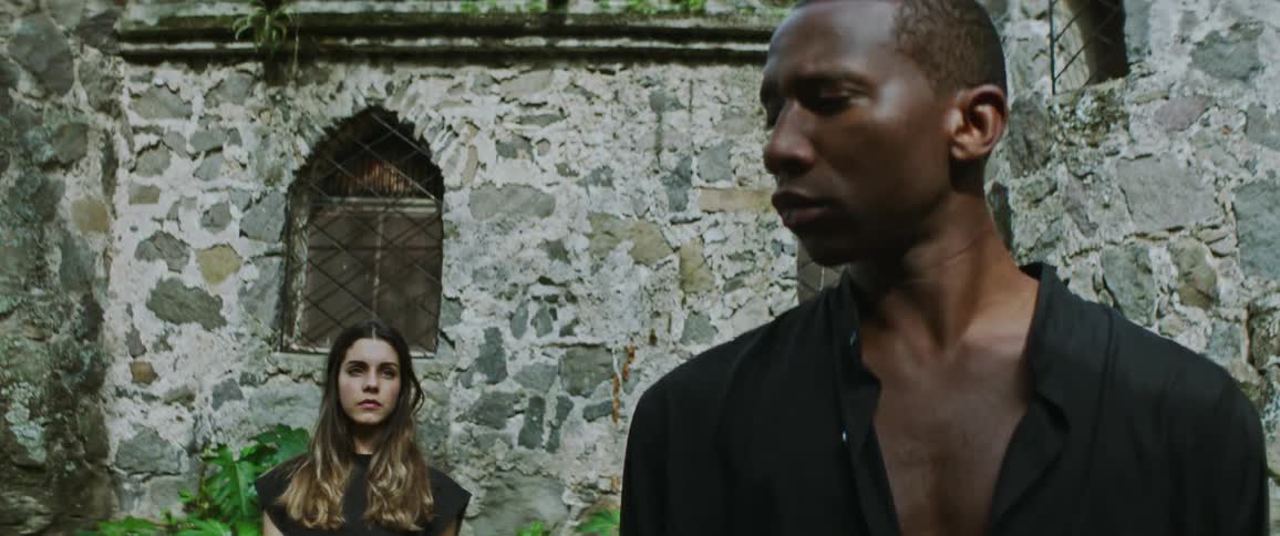 A man standing in front of a stone building turns his head to look at the woman behind him