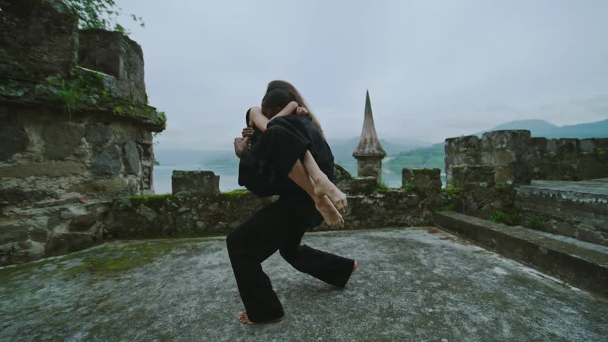 Male dancer with black shirt and pants holds woman wearing black dress in his arms on top of castle tower
