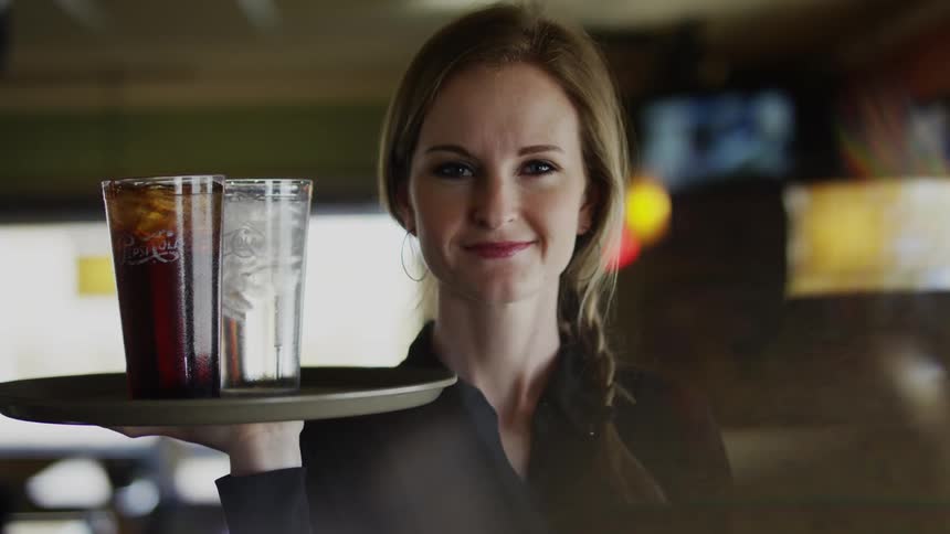 A waitress stands smiling while holding a tray up with two drinks on it