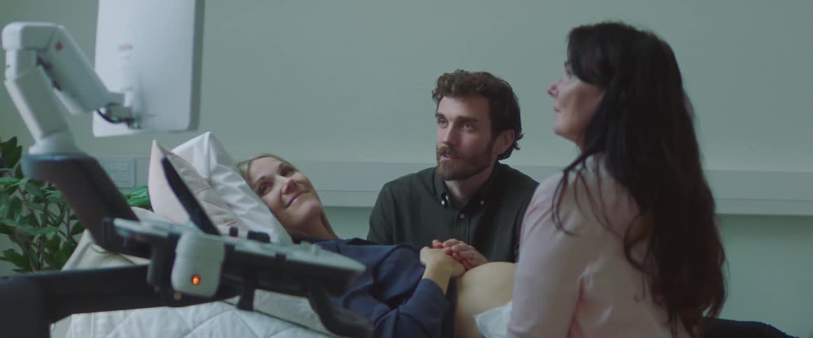 A medical technician is performing an examination on a pregnant woman as they smile and look at a monitor