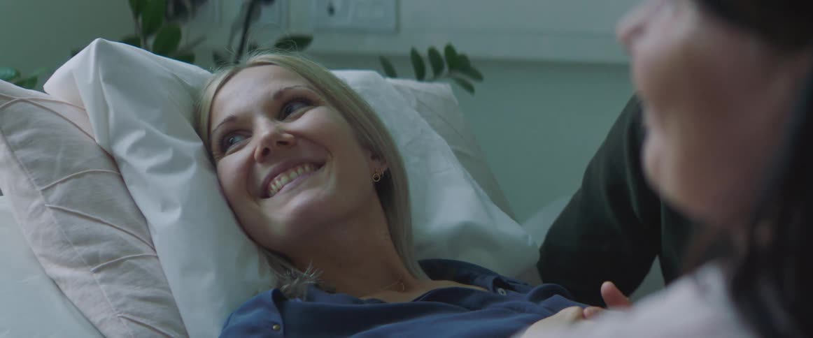 A woman lying in a bed holding her husbands hand listens to a nurse and then smiles and turns to her husband