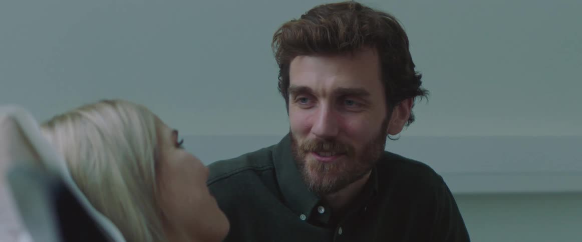 A man sits next to a woman who is propped up in bed and they smile at one another and talk
