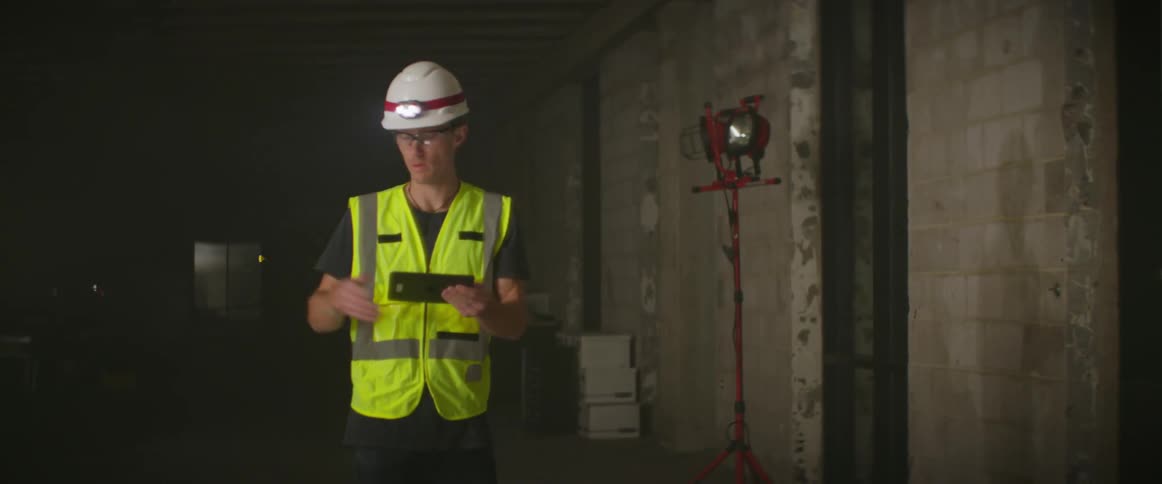Construction worker wearing bright yellow vest and white hardhat supervising a construction site at night