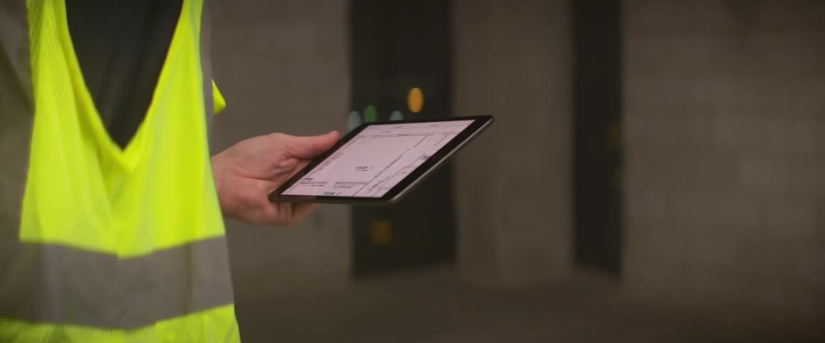 Construction worker wearing bright yellow vest and white hardhat supervising a construction site at night with a tablet