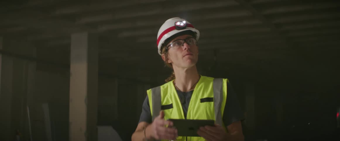 A man wearing a hardhat and safety vest holding a tablet points at beams and ceiling