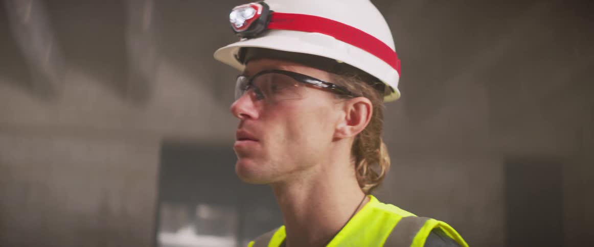 A man wearing a hardhat and safety goggles points up at the ceiling as he talks