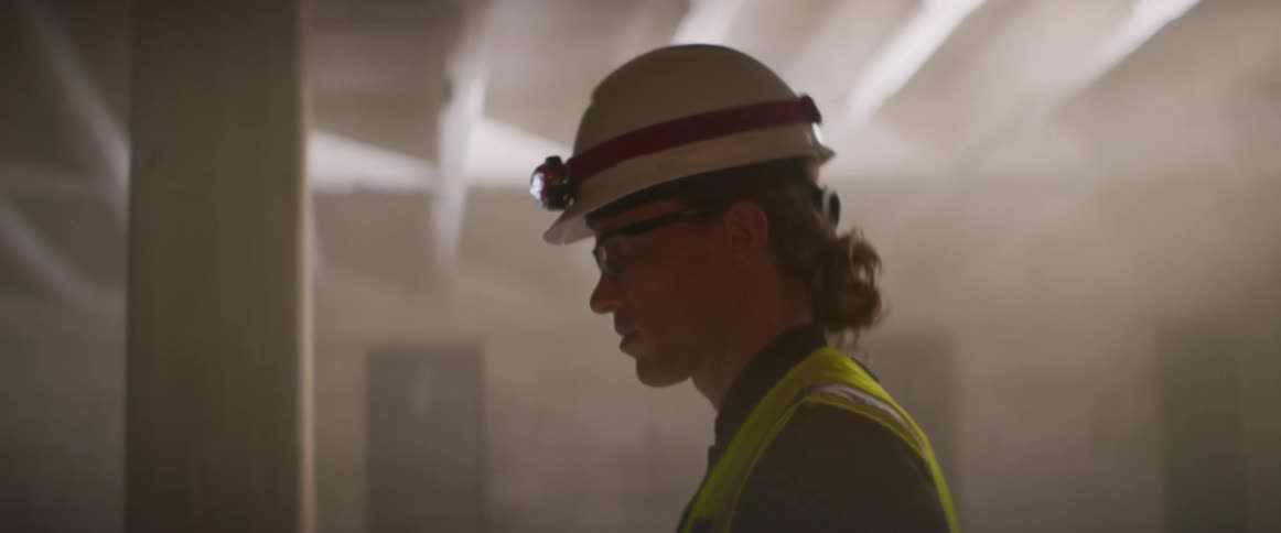A man points up at the ceiling beams as he walks around wearing a hardhat with head lamp