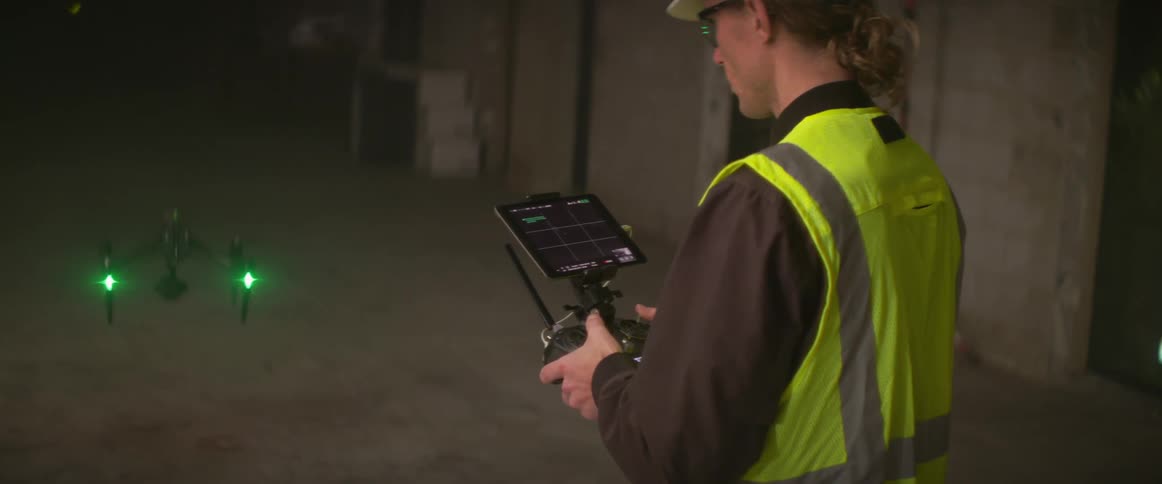 Worker wearing a safety vest and a hard hat stands in building controlling a drone